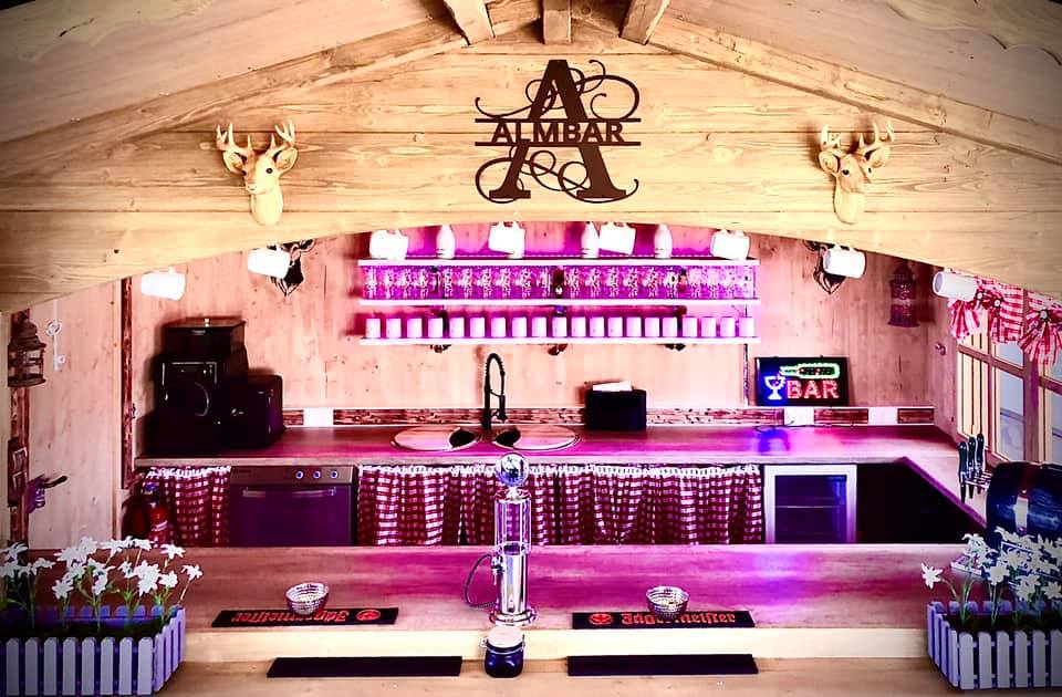Eine mobile Bar im rustikaler Holzhüttenoptik, mit Theke und Spülbecken. Im Wandregal stehen Gläser und Krüge.