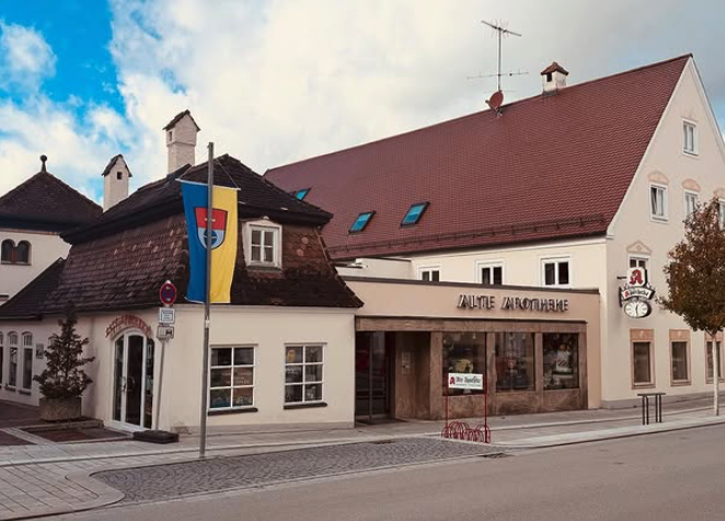 Eine Gebäude in beigem Farbton, bestehend aus einem Altbau und einem Neubau, an dem eine Uhr und ein Apothekenschild angebracht ist.