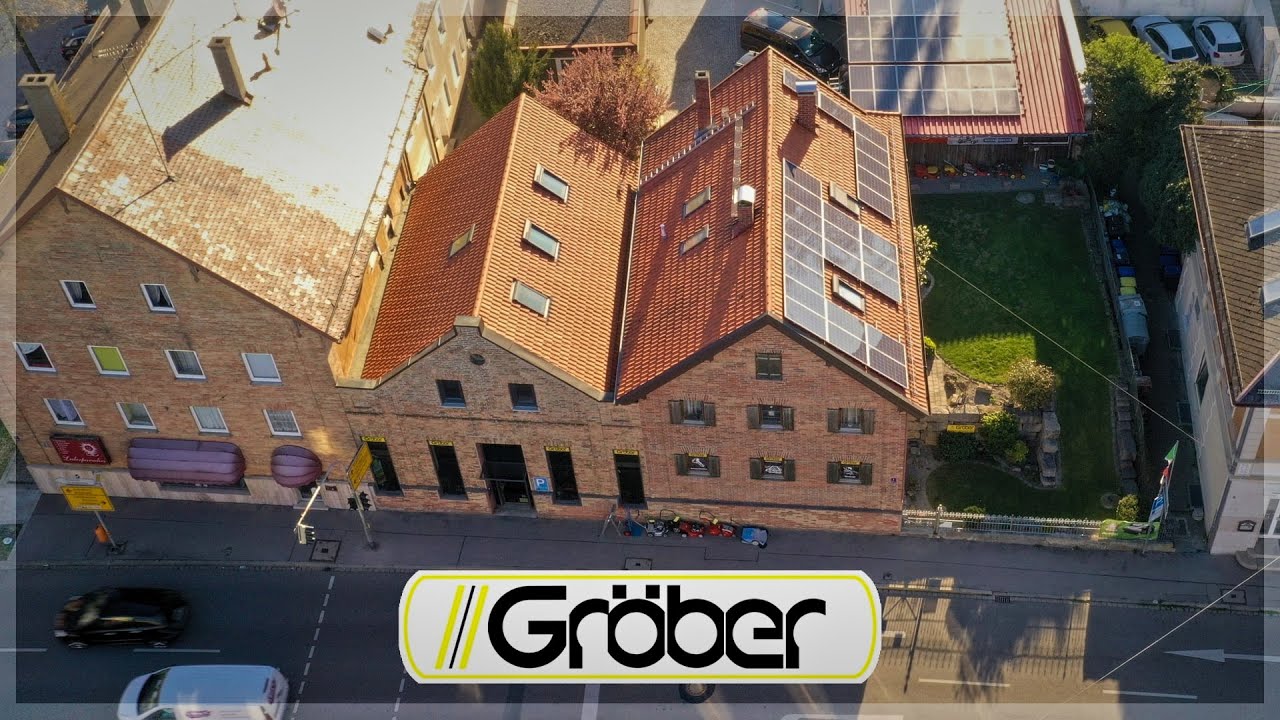 Luftansicht eines Backsteingebäudes mit Schaufenstern und Garten. Davor stehen Rasenmäher.