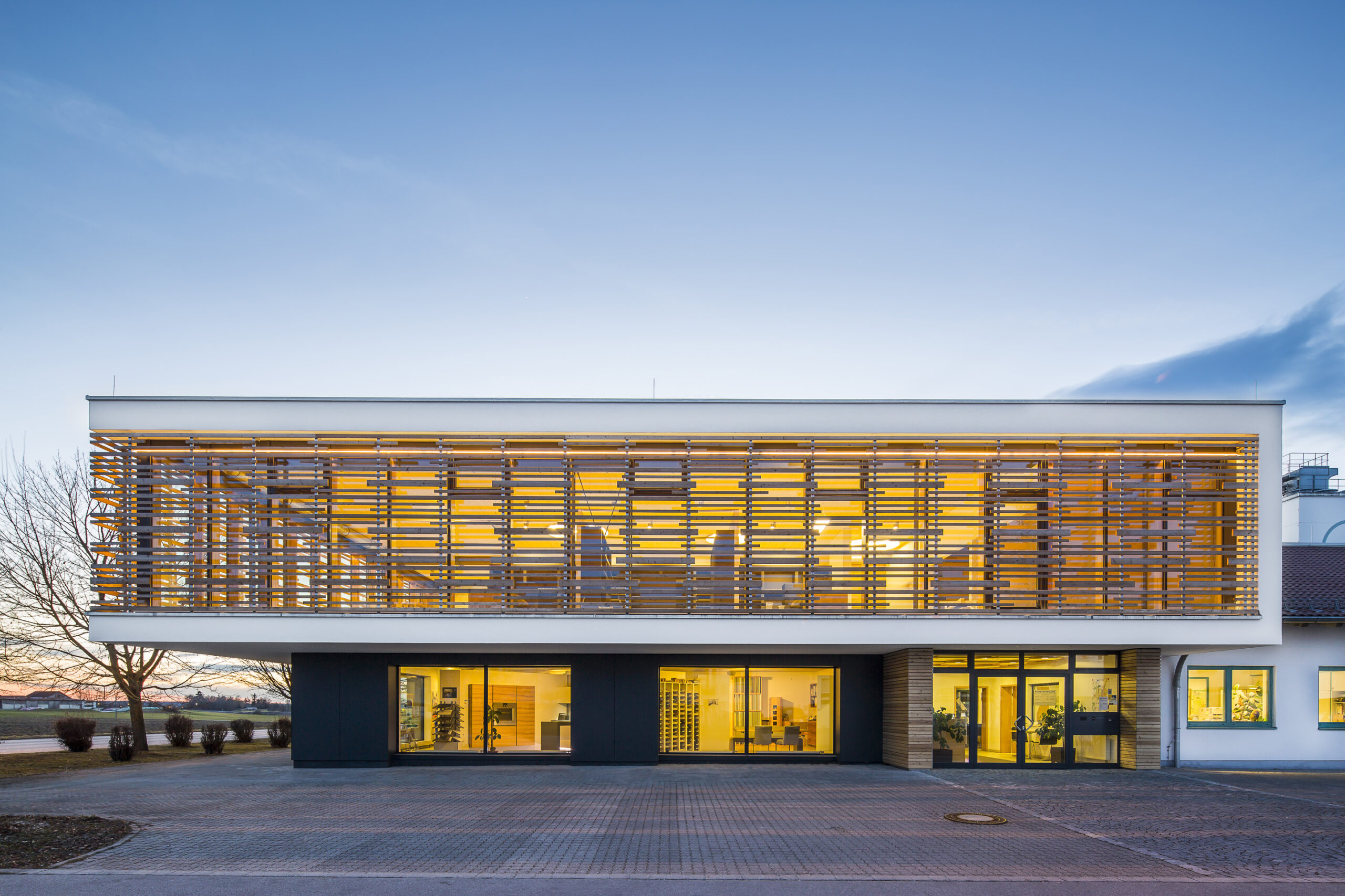 Modernes Gebäude im Bauhausstil mit großen, von innen beleuchteten Fenster. Im Hintergrund Himmel in der Abenddämmerung.