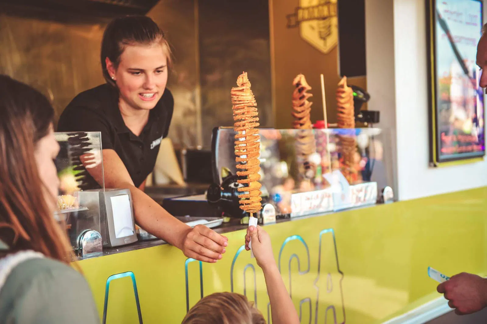 Eine junge Frau, die einem Kind eine auf einen Spieß abgedrehte Spiralkartoffel reicht. Sie lehnt aus dem Fenster bzw. der Anreiche eines Foodtrucks.