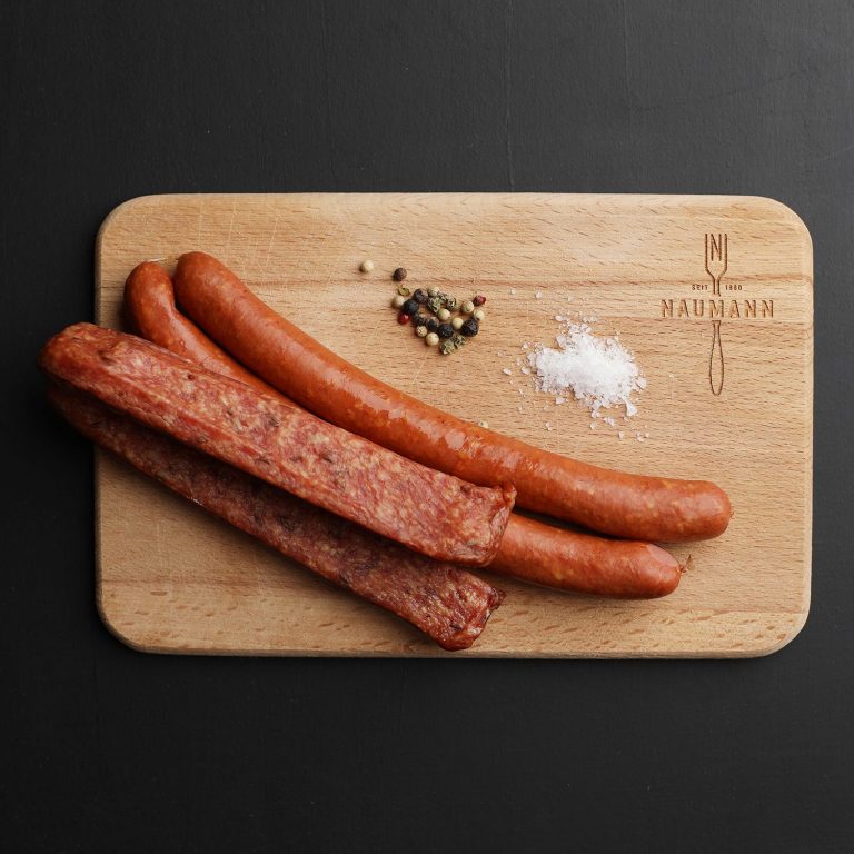 Ein Holzbrett auf schwarzem Hintergrund mit zwei paar Würsten darauf und Salz und Pfeffer.