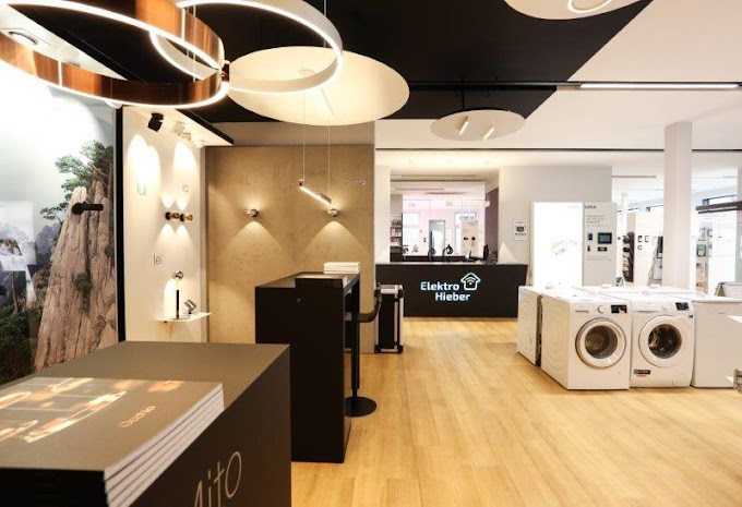 Innenansicht eines modernen Showroom mit Holzboden und schwarzen Paneelen. Ausgestellt sind verschiedene Elektrogeräte, unter anderem Waschmaschinen.