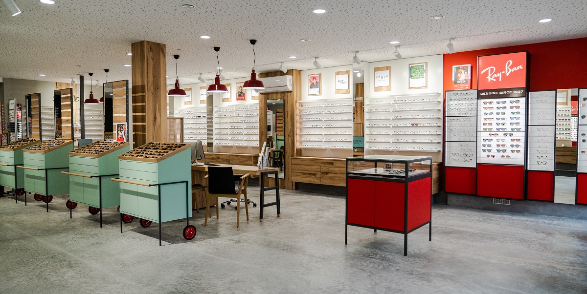 Ein moderner Verkaufsraum ihm grauem Boden und Regalen aus Holz und Glas, mit abgesetzten Roten und mintgrünen Akzenten. In den Regalen sind Brillen ausgestellt, man sieht außerdem einen Tisch mit Stühlen und einen Spiegel.