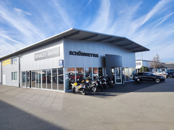 Eine Gewerbehalle auf betonierter Fläche, große Schaufensterflächen und Motorräder und Autos außen aufgestellt.