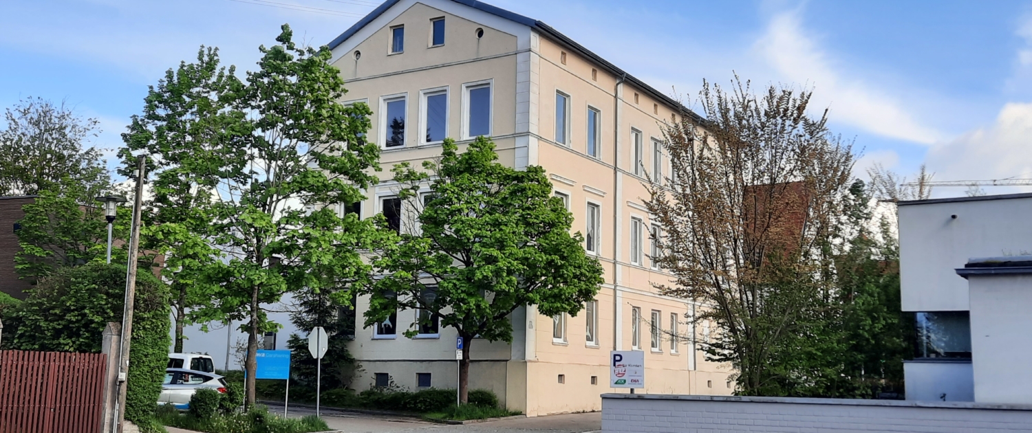 Modernisierter Altbau mit gelber Fassade, umrahmt von Bäumen vor blauem Himmel.