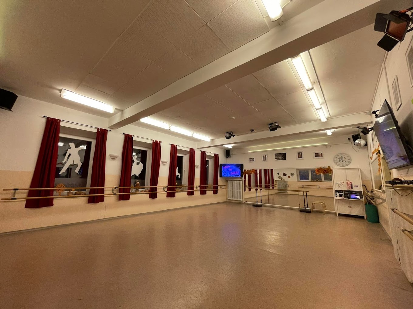 Ein großer Raum mit Parkettboden, Fenstern mit roten Vorhängen, einem großen Spielgel und Ballettstangen an der Wand.
