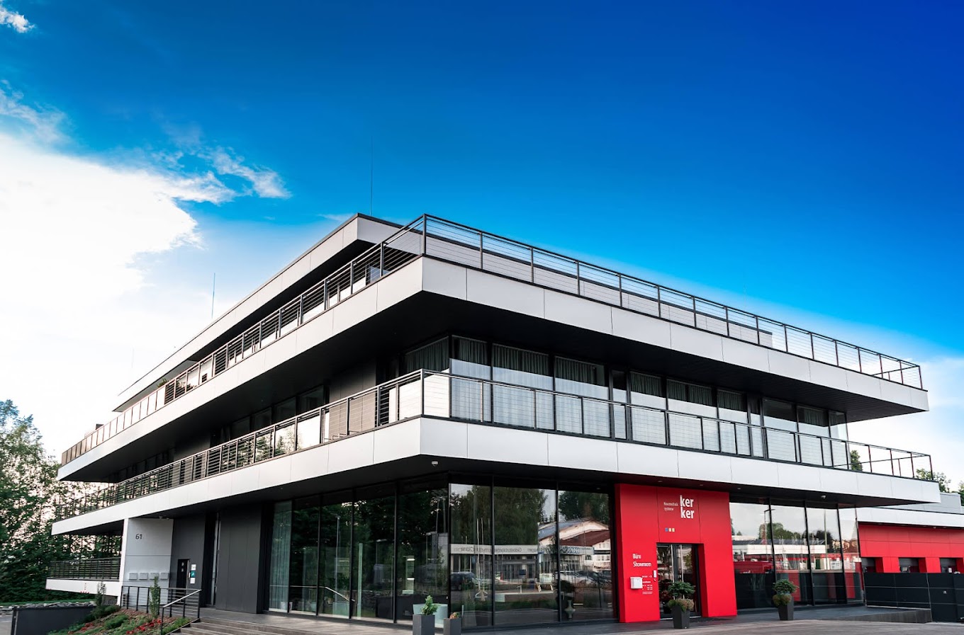 Ein moderner Flachdachgebäude mit Terassenartiger Stockwerksanordnung und Glasfassade bzw. Glasbalustraden. Abgesetzter Roter Eingang.