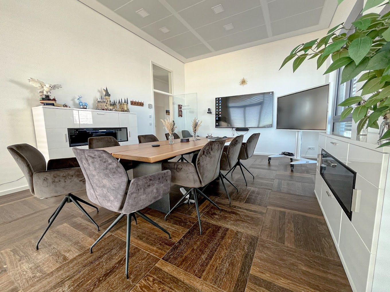 Konferenzraum mit dunklem Parkett und weißen Wänden. In der Mitte ein Holztisch, darum herum grau-braune Velours-Stühle. Zwei große Bildschirme an der Stirnseite.