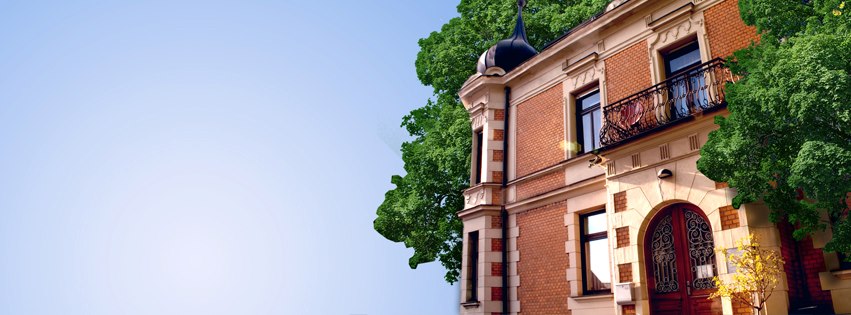 Außenansicht einer Stadtvilla aus Backstein im Jugendstil mit geschwungenem Balkongeländer und eleganter Flügeltür aus Holz als Eingang vor blauem Himmel.