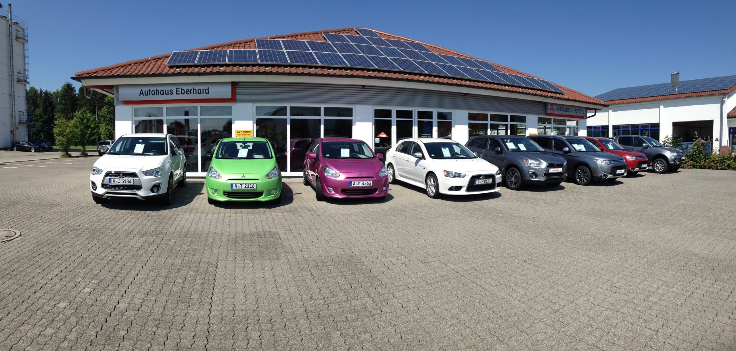 Eine reihe PkW mit der Stoßstange zum Betrachter aufgestellt von einem Autohaus. Die Sonne scheint, der Himmel ist blau und der Lack glänzt.