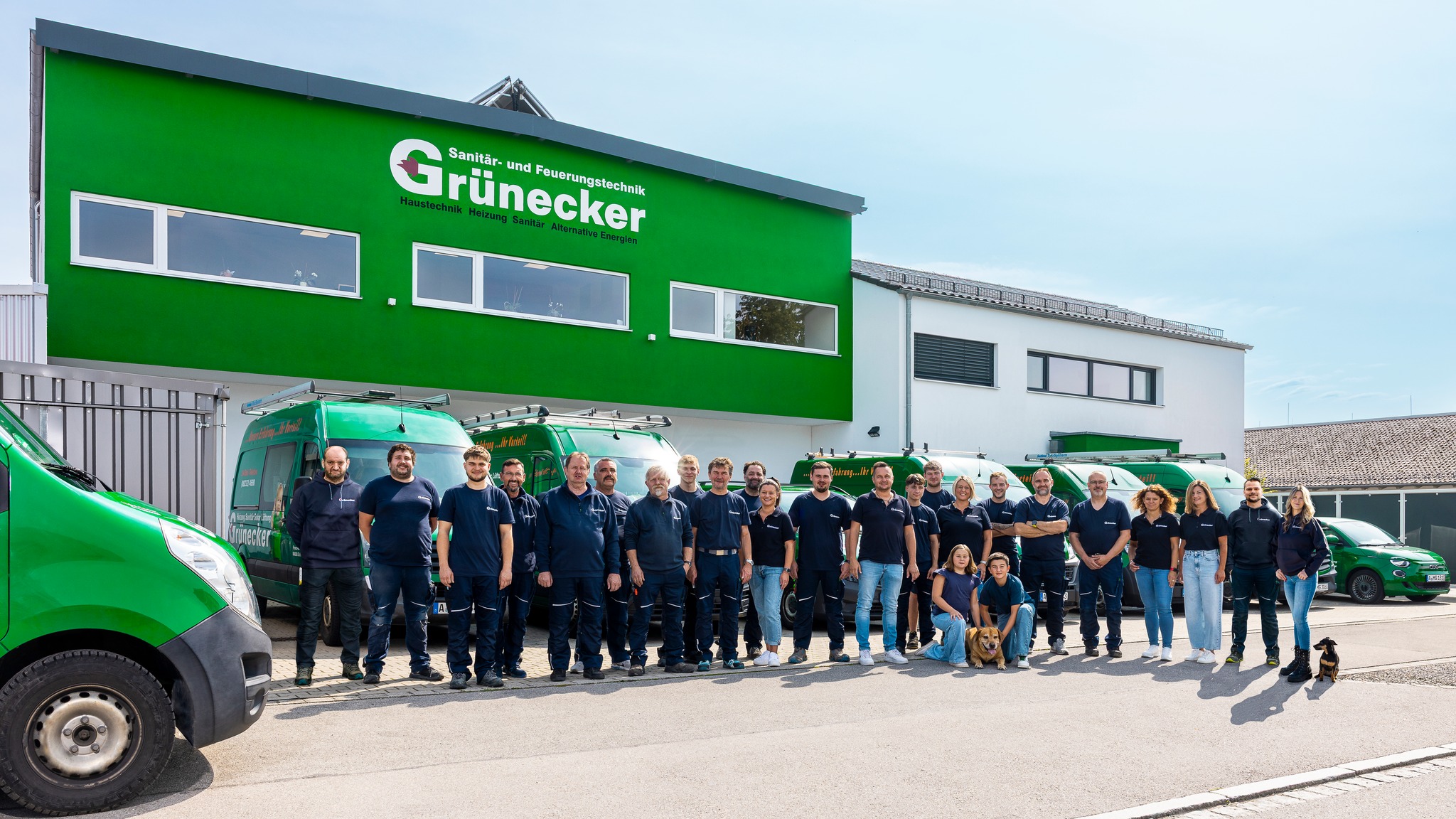 Ein modernes Gebäude mit grasgrüner Fassade, davor Transporter im selben Grün und eine Gruppe Menschen in dunkelblauer Arbeitskleidung, sowie zwei Hunde, die in die Kamera schauen.