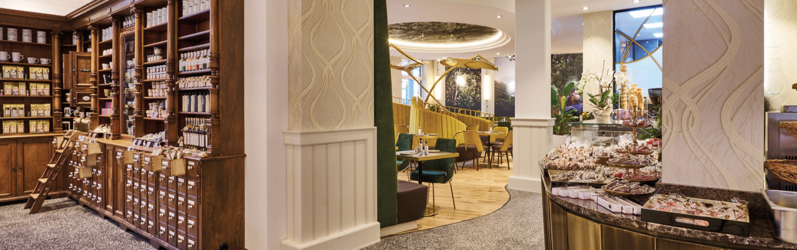 Blick in den Cafébereich mit stilvollem Mobiliar und goldenen Elementen. Teilweise Ladenbereich mit alten Apothekerschränken aus Holz.