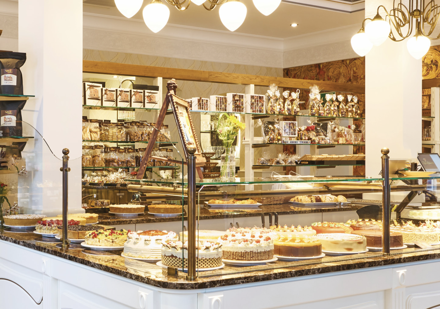 Theke mit braunen und goldenen Elementen. In der Theke befinden sich kunstvolle Torten, Gebäck, Pralinen und Schokoladen.