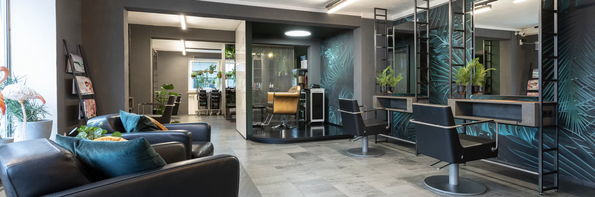 Ein moderner Raum mit Friseurstühlen, und Spiegel in dunklem Metalllook. An der Wand eine schwarz-grüne Tapete mit Duschungeloptik. Wartebereich mit schwarzen Ledersesseln.