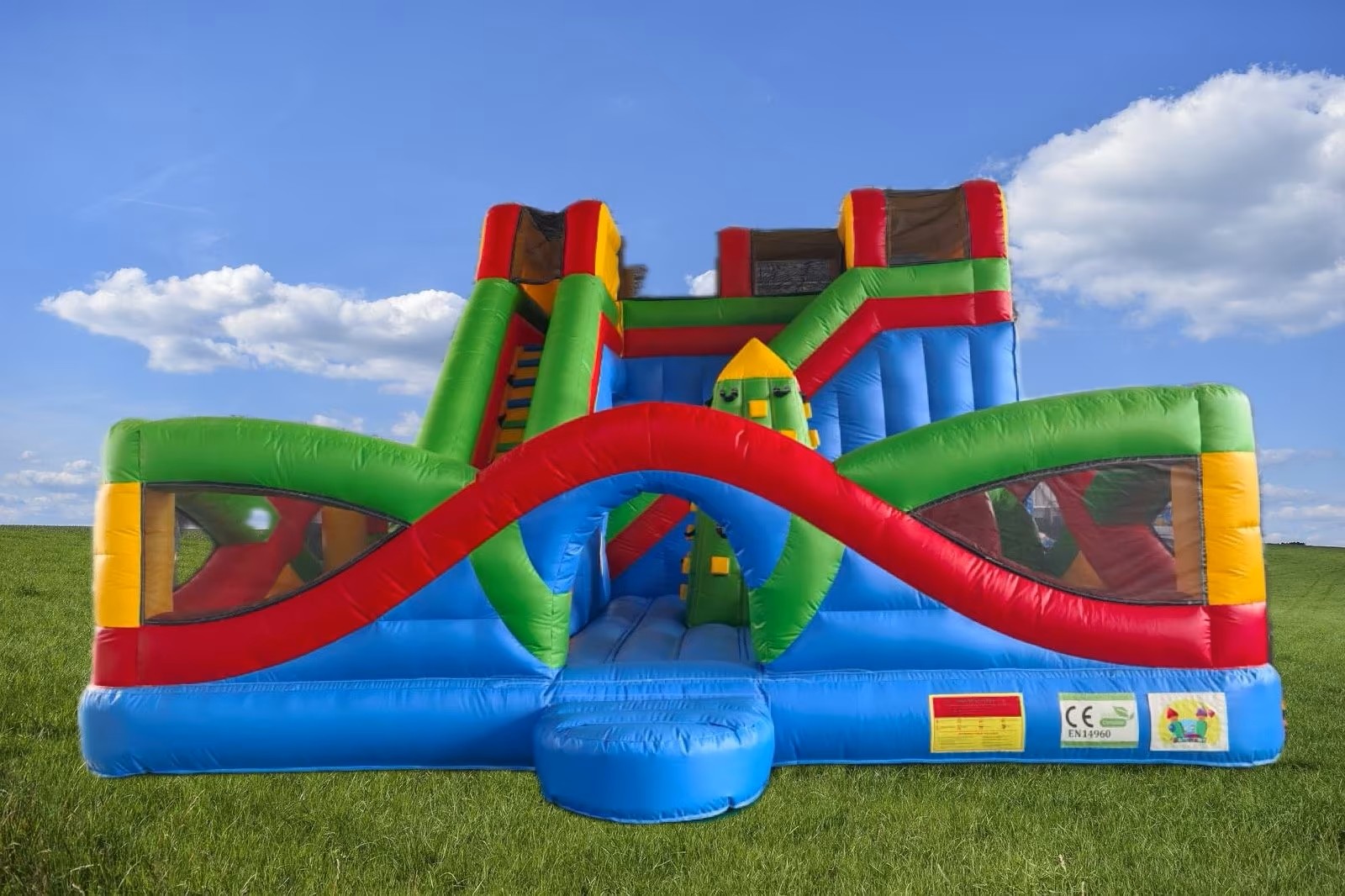 Eine Wiese mit blauem Himmel, darauf eine große Hüpfburg in Primärfarben mit Rutsche.