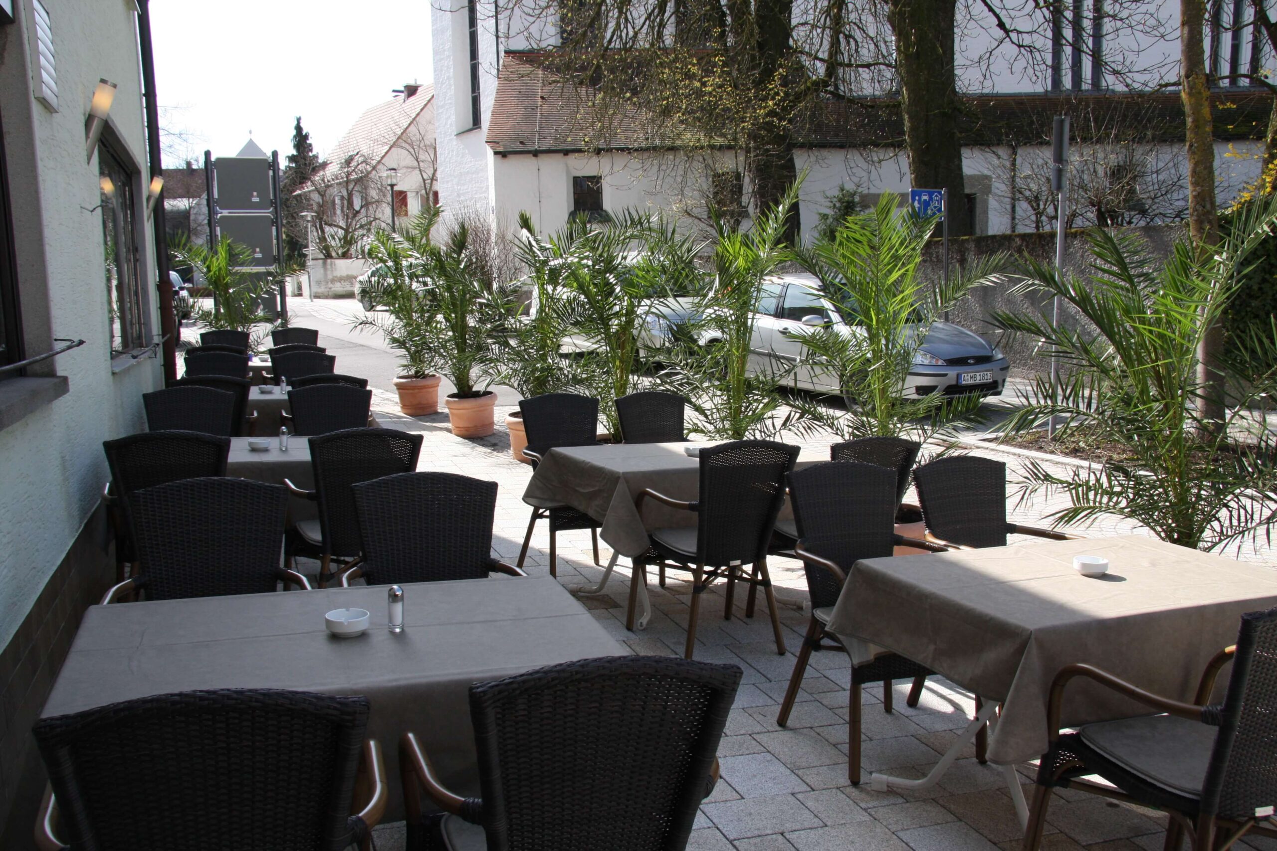 Eine Reihe schlicht gedeckter Tische und Stühle auf einer Terrasse umringt mit kleinen Palmen, im Hintergrund eine Kirche.