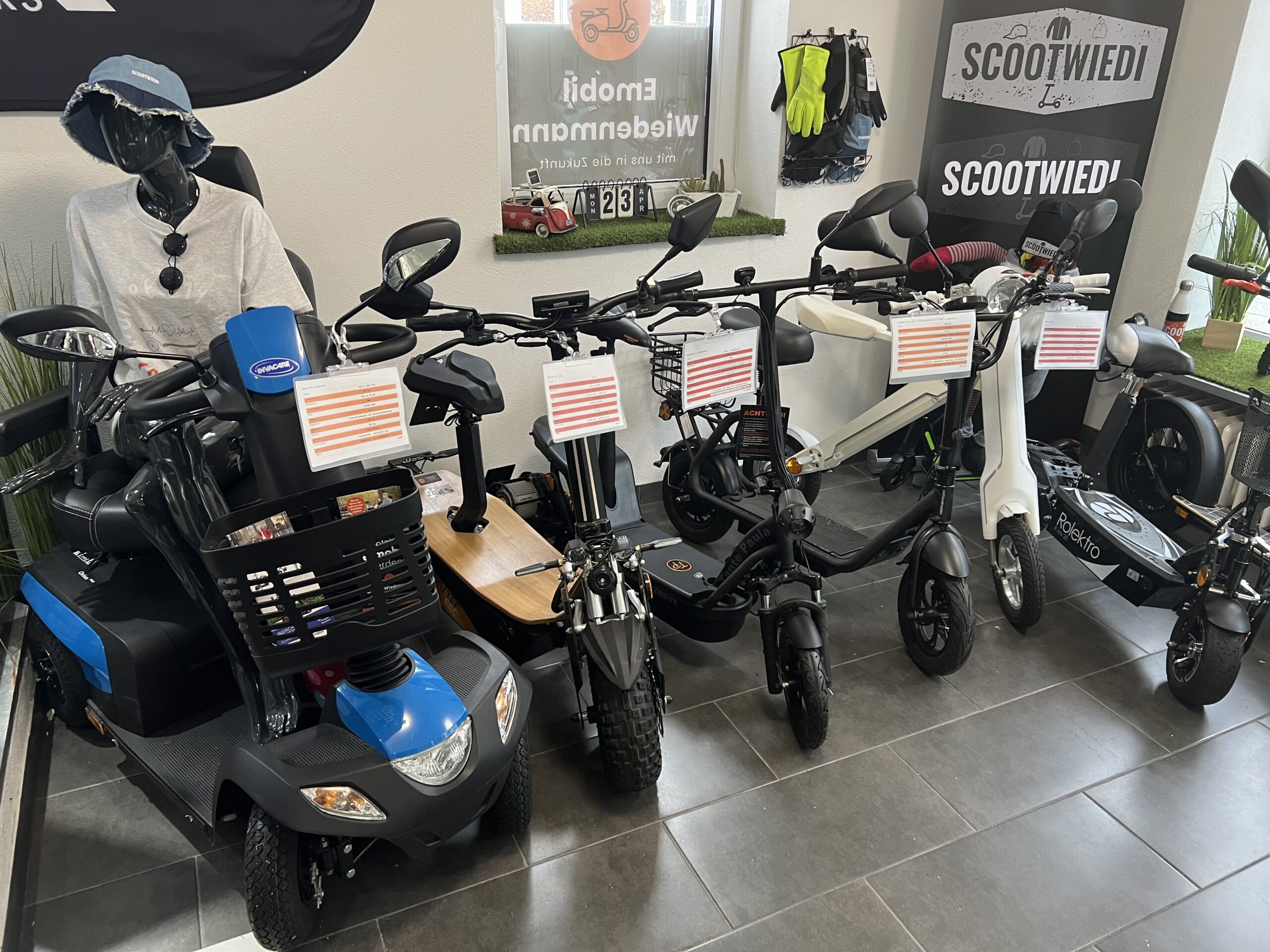 Ansicht einer Reihe Fahrzeuge, ausgestellt im Innenbereich, darunter ein Seniorenmobil, E-Scooter und E-Bikes.