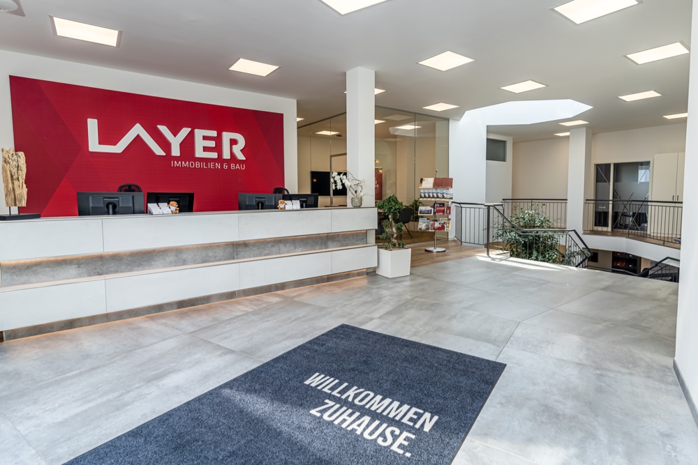 Heller Innenraum mit Steinboden und quadratischen Oberlichtern. Ein langer Empfangstresen mit Computerbildschirmen dahinter, ein Gang und eine Treppe, die abwärts führt.