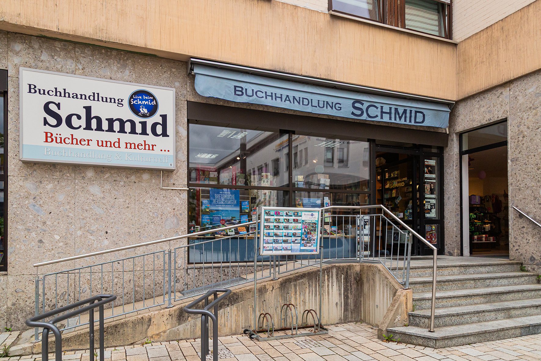 Außenansicht einer Buchhandlung mit Büchern und Geschenkartikeln im Schaufenster. Treppen und Rampe als Aufgang, blaue Markise über Schaufenster und Eingang.