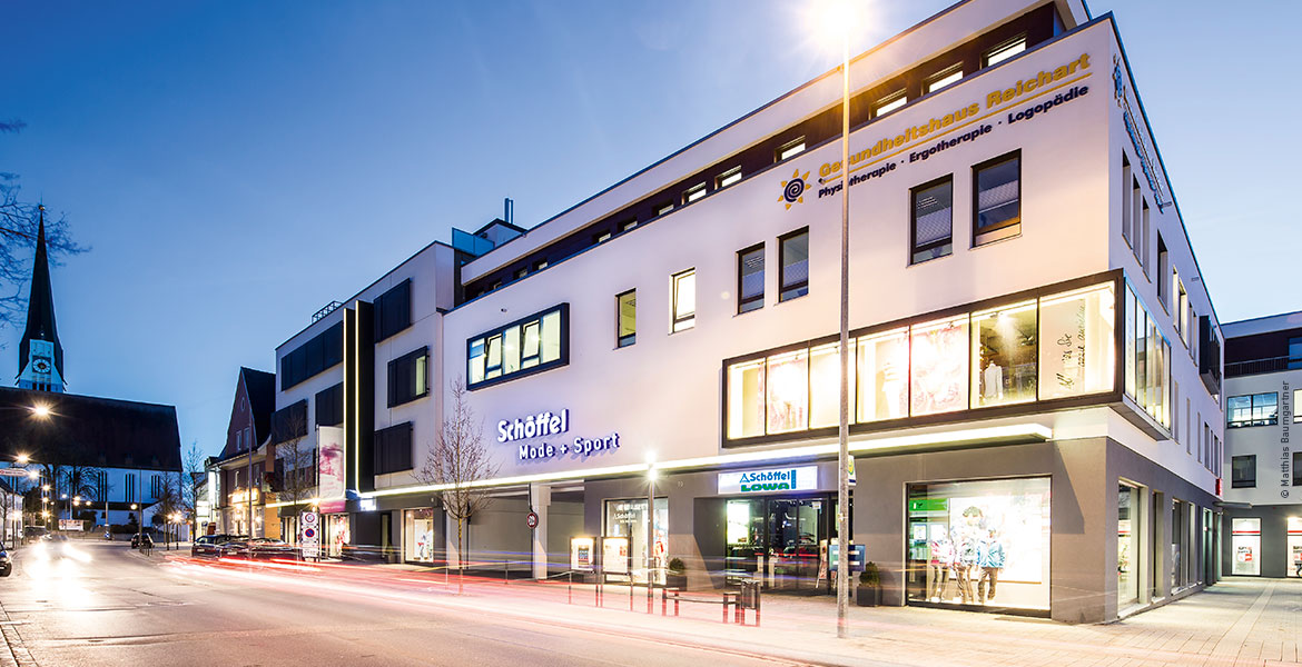 Ein modernes Gebäude mit weißer Fassade an einer Straße bei Nacht. Beleuchtete Schaufenster zeigen Mode.