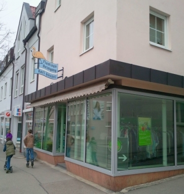 Außenansicht eines Gebäudes mit großer Fensterfront im Erdgeschoss. Durch die Fenster sind ordentlich aufgehängte Kleidungsstücke zu erkennen, außen ist ein Schild befestigt, auf dem Flamingo Reinigung Karl Göppel steht.