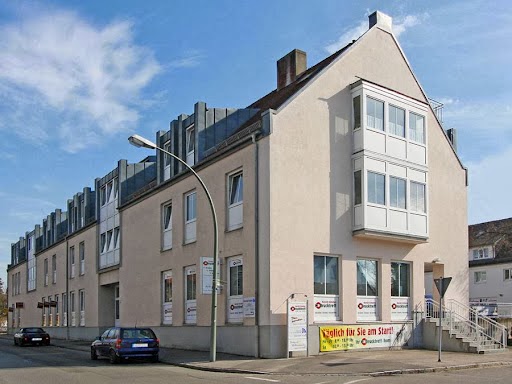 Außenansicht eines modernen, beigen Gebäude. In das Gebäude führt eine Treppe. Schaufenster sind mit Werbung beklebt.