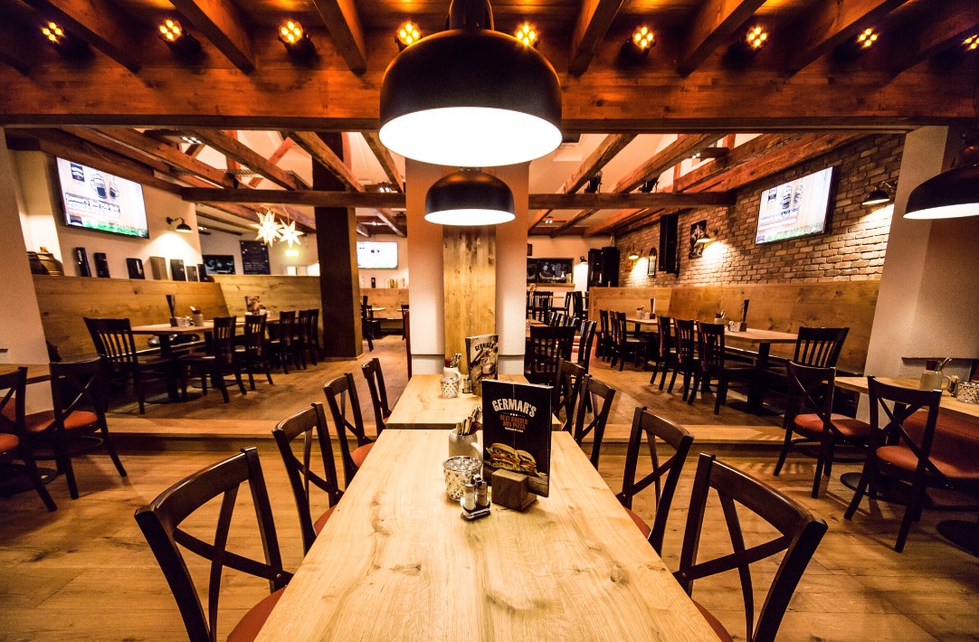 Innenansicht eines Restaurants, eingerichtet im rustikalen Stil mit viel Holz, offenen Balken und Backstein. Zentral im Bild ein großer Holztisch mit Speisekarte, oben na der Decke hängen große schwarze Leuchten.