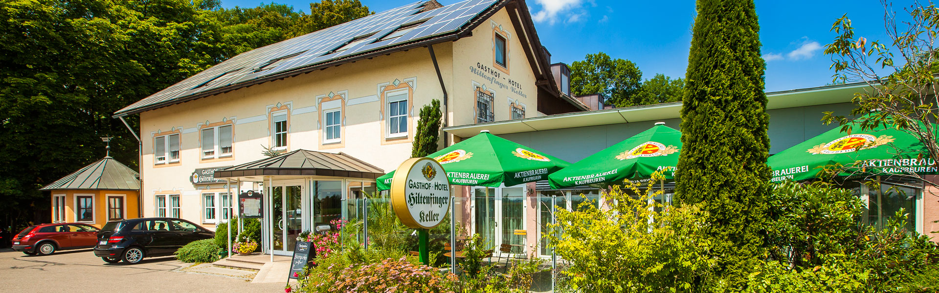Ein Gebäude mit Giebeldach mit traditionell gestrichener Fassade umgeben von Bäumen und Büschen. Gründe Sonnenschirme mit Brauereilogo und Stühle und Tische auf der Terrasse.