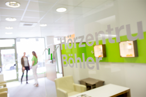 Eine Glastür mit dem Aufdruck Hörzentrum Böhler, im Hintergrund außer Fokus ein weißgrün gestrichener Eingangsbereich, eine Eingangstüre und zwei Menschen, die sich unterhalten.