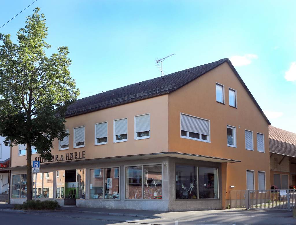 Ein ockerfarbenes Gebäude aus den 70ern mit Schaufensterfront im Erdgeschoss und angeschlossener Scheune. Im Vordergrund ein Baum.