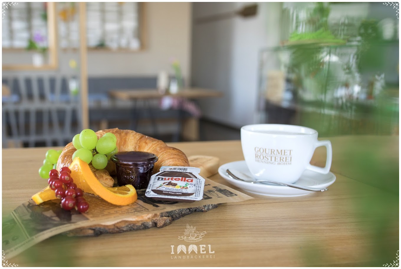Eine weiße Kaffeetasse, ein Frühstück mit Obst und Croissant auf einem Holztisch in einem Café.