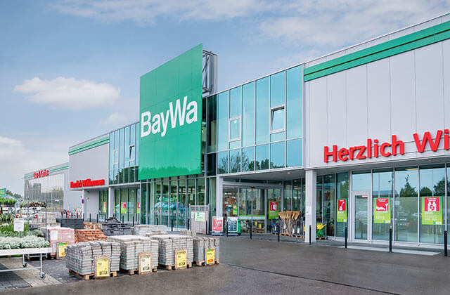 Ein modernes Gebäude mit grau-grüner Verkleidung und Glasfront im Eingangsbereich. Pflanzen, Baumaterial und anderen Waren sind im Außenbereich ausgestellt.