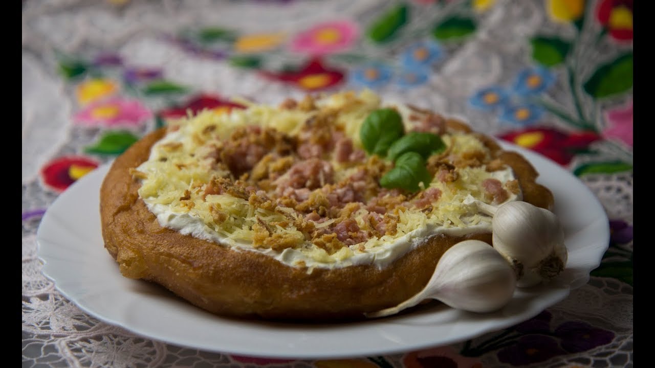 Ein frittiertes Mangos Küchle auf einem weißen Teller. Auf dem Langos ist Schwand, Käse und Rostwiebeln, zwei Knoblauchzehen liegen auf dem Teller als Deko.