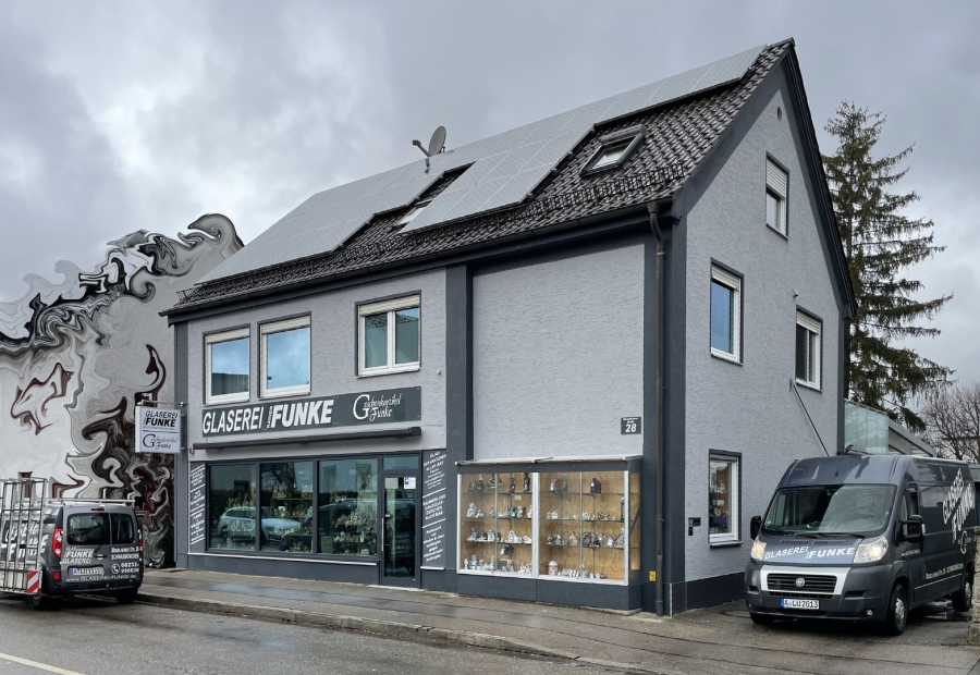 Außenansicht eines grauen Gebäudes mit Schaufenster im Erdgeschoss und Glasschaukasten neben der Eingangstür. In den Schaufenstern erkennt man Dekorationsartikel, in der Einfahrt steht ein grauer Transporter.