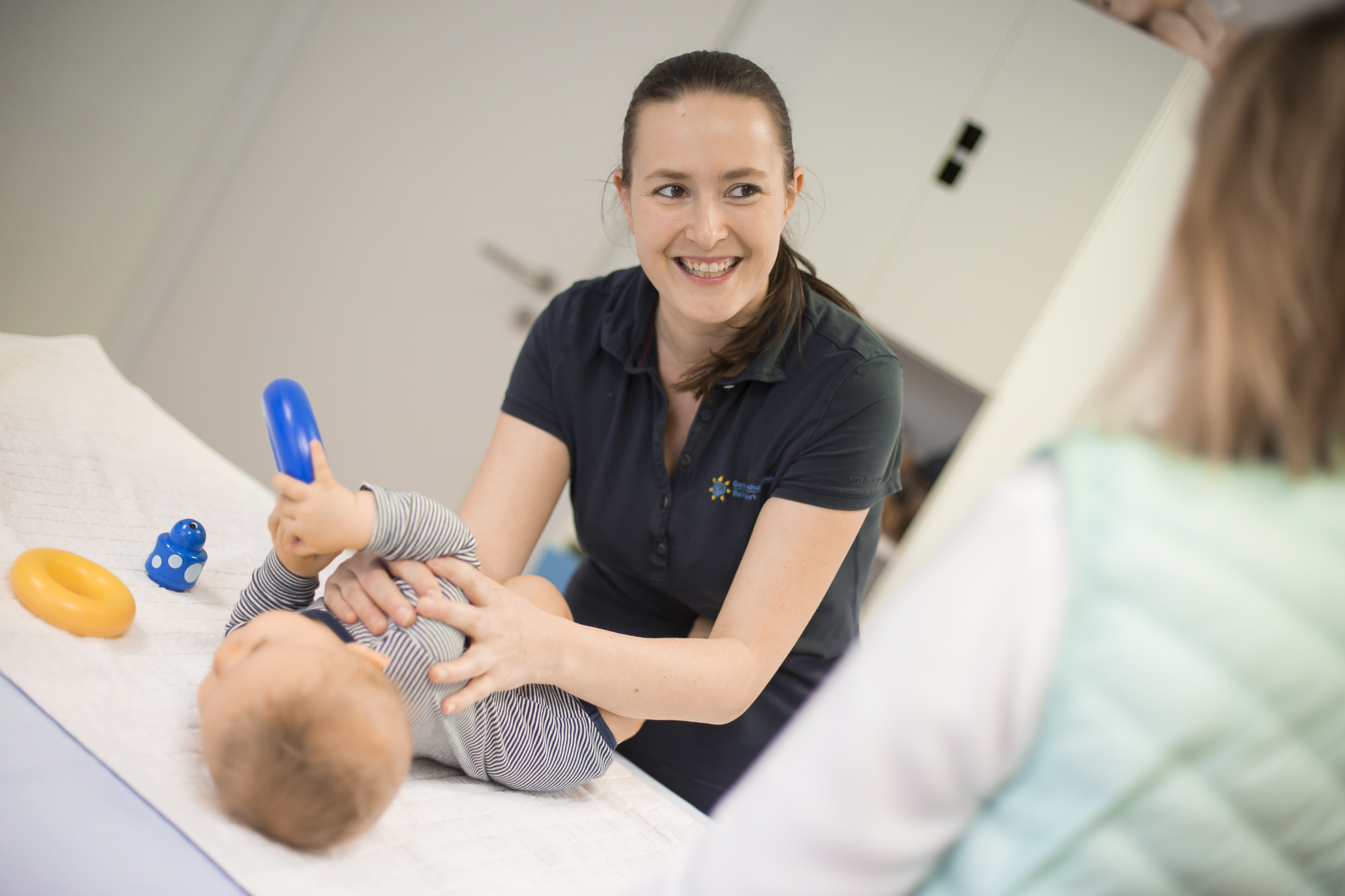 Eine junge Frau über einen Säugling auf einem Behandlungstisch gebeugt. Sie lächelt einer Mutter zu, die mit dem Rücken zur Kamera am Bildrand zu erkennen ist.