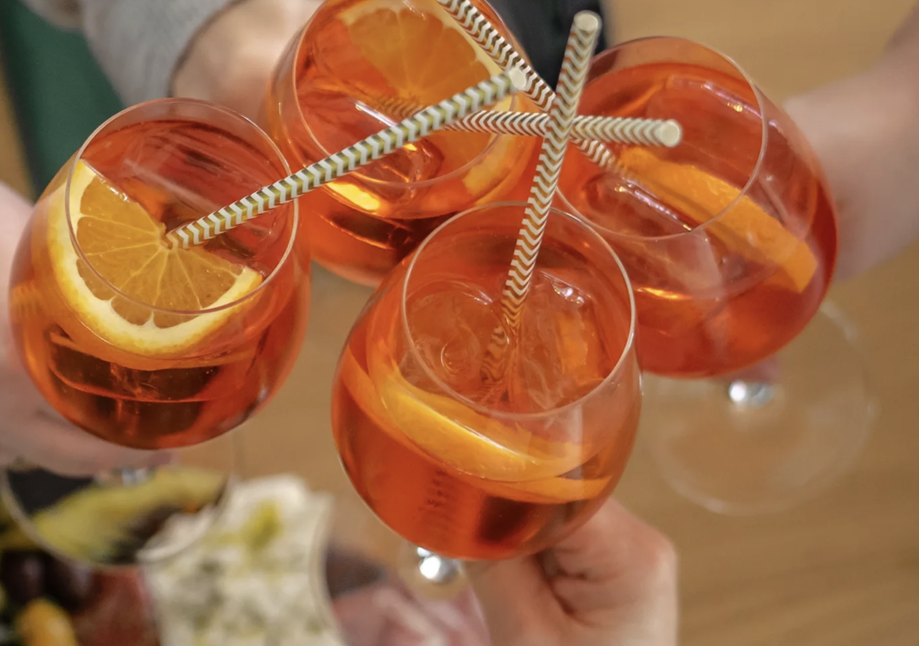 Vier Aperol-Spritz mit Orangenscheibe und Eiswürfeln und Strohhalm werden von Händen gehalten. Es wird angestoßen.