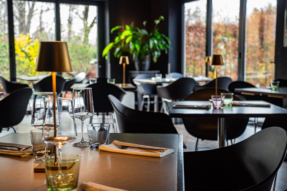 Ein dunkel gestrichener Raum mit großen Fenster. Schlichte dunkle Tische und schwarze Stühle, elegantes Flair und gedeckte Tische mit Weingläsern.
