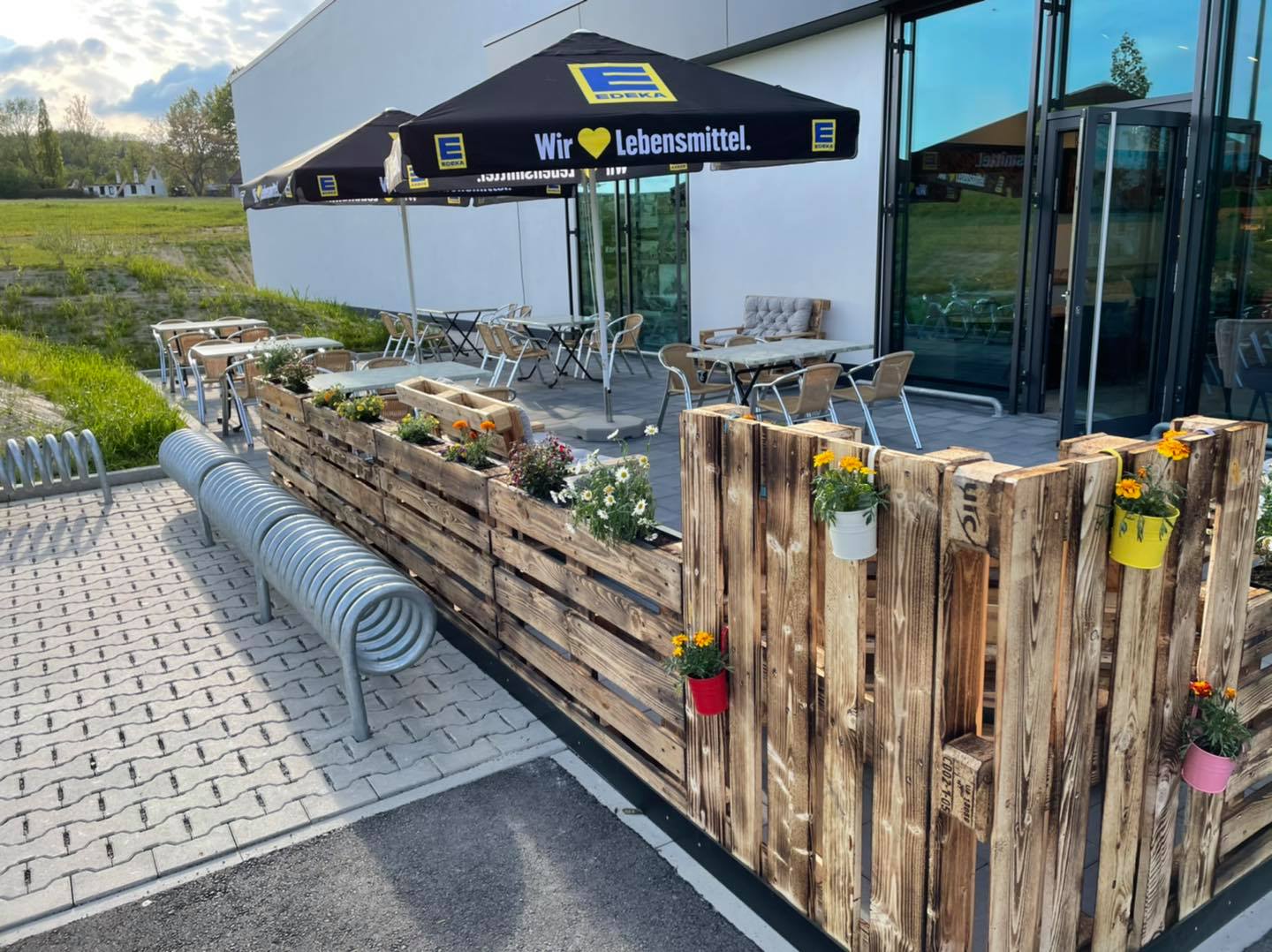 Ein mit Holzpaletten eingezäunter Bistro Bereich mit Stühlen und Tischen, Fahrradständer aus Metall im Vordergrund