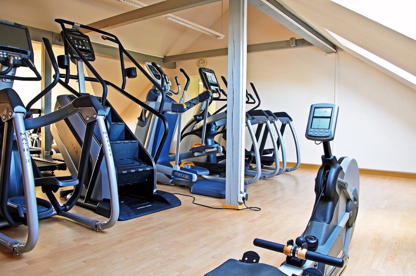 Heller Raum mit Holzboden und Dachschrägen, darin Fitnessgeräte wie Crosstrainer oder Fahrradergometer.